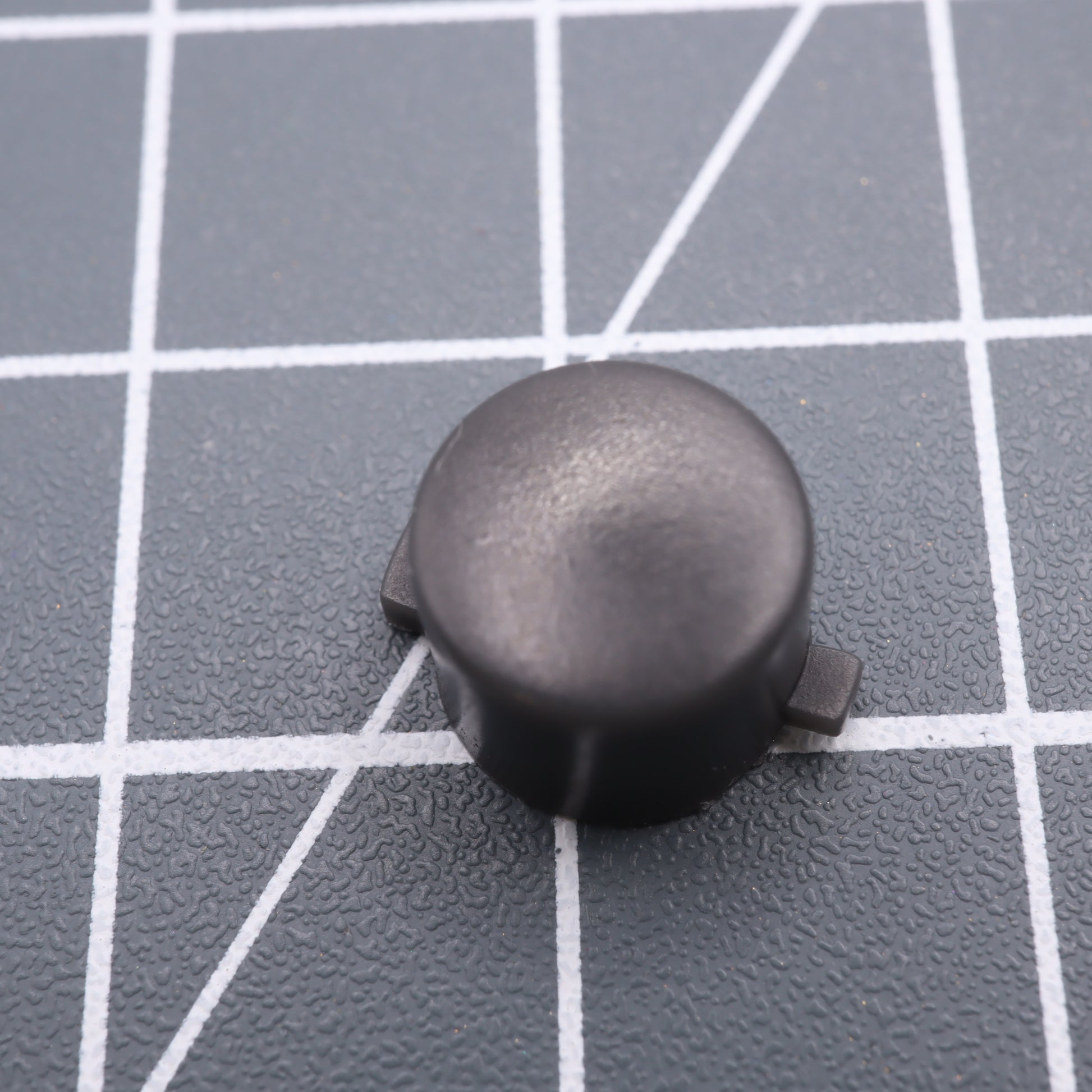 A DMG Play it Loud Style custom black round resin button with side tabs from an Analogue Pocket is on a gray grid surface.