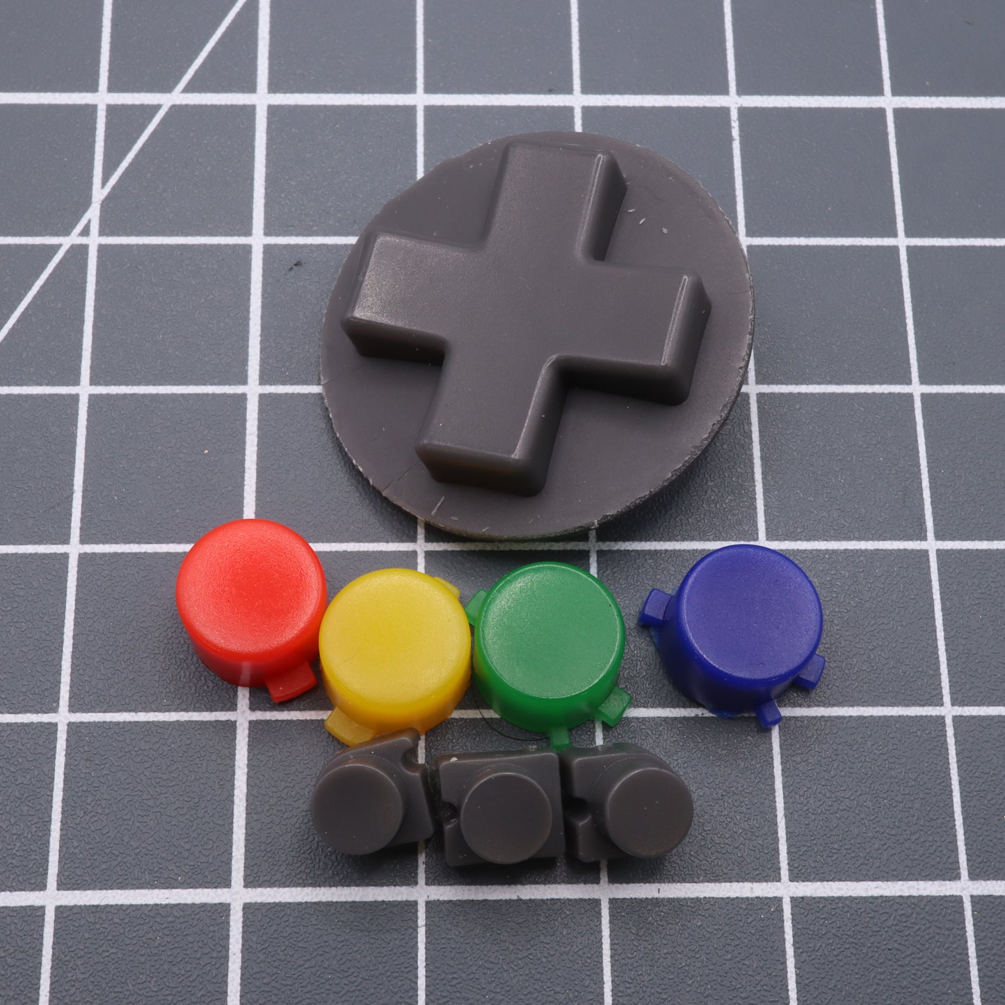 The Analogue Pocket - Custom Button - SNES Style set includes a black directional pad with red, yellow, green, blue, and black buttons, made using custom-cast molds and stylishly displayed on a gray grid surface.