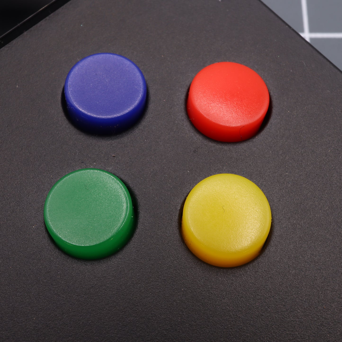 Close-up of four vibrant buttons on a black surface, crafted with custom-cast molds in blue, red, green, and yellow—reflecting the iconic hues of Analogue Pocket's SNES Style Custom Buttons.