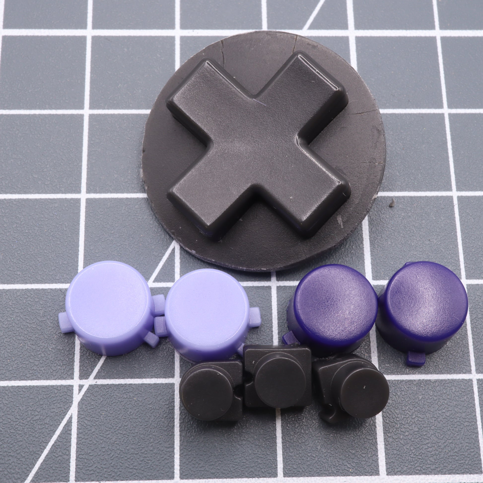 A set of circular and cross-shaped game controller buttons in purple, black, and dark blue, crafted from custom-cast molds on a grid-patterned surface for the Analogue Pocket - Custom Button - Famicon Style.