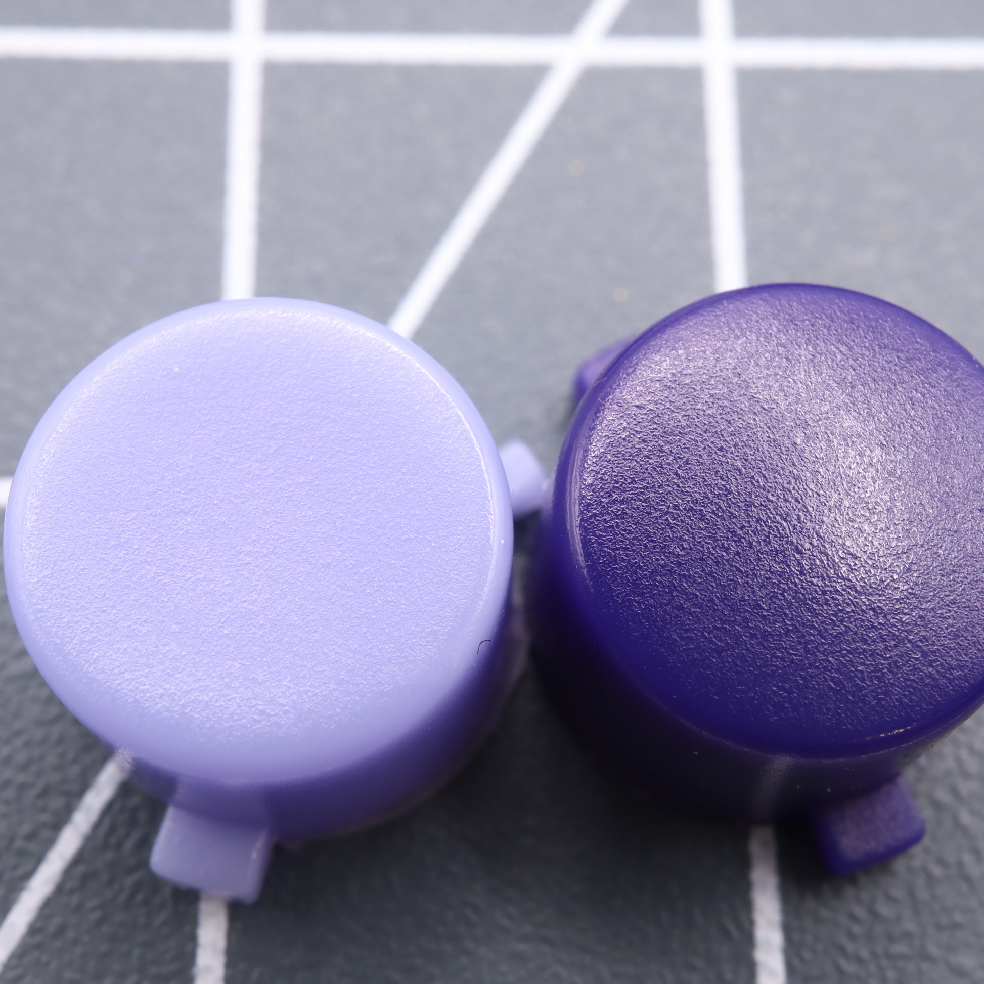Two round, purple plastic caps rest on a textured gray surface with white grid lines. These custom-cast buttons, inspired by Famicom style from Analogue Pocket - Custom Button collection, perfectly merge retro aesthetics with modern craftsmanship.