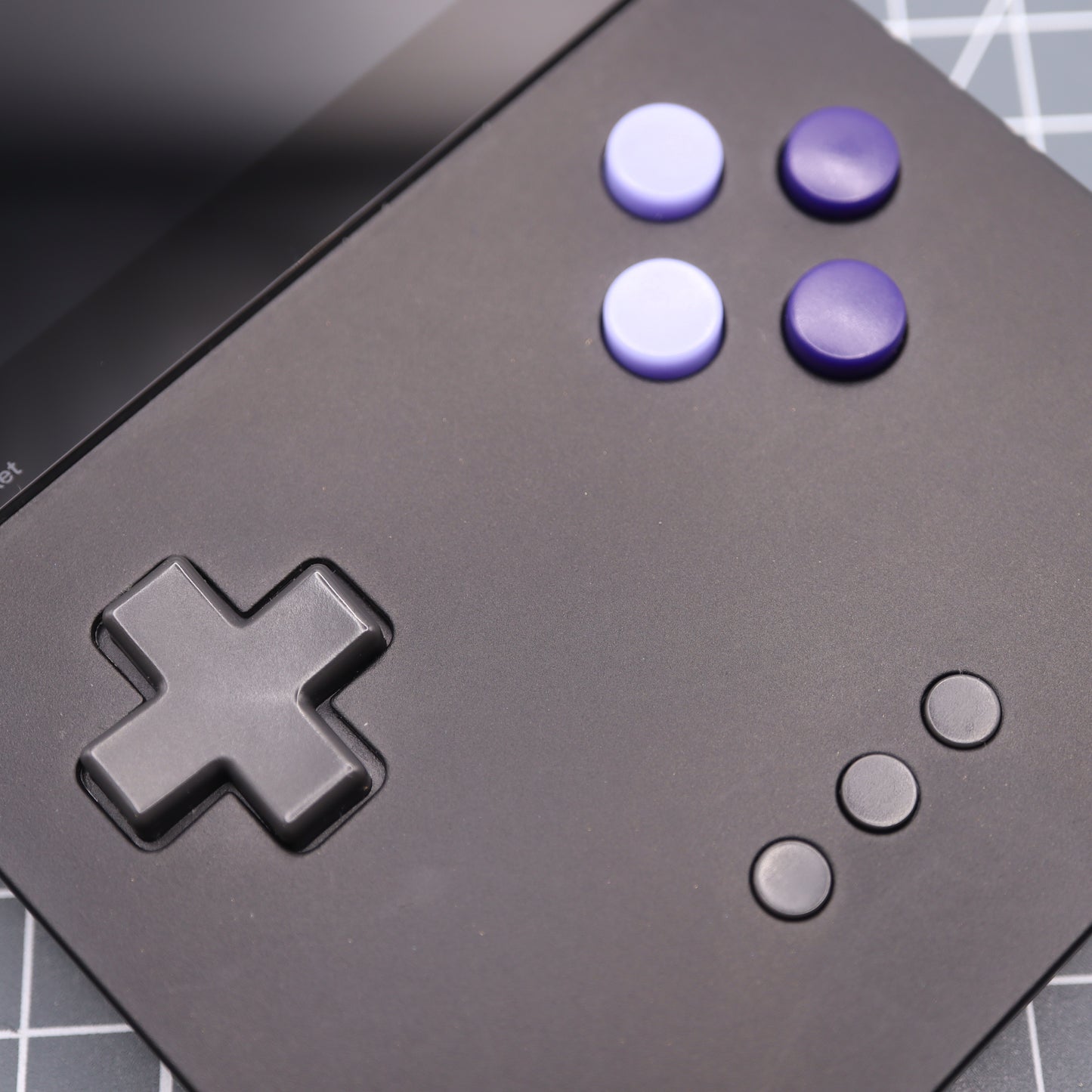 A close-up of the Analogue Pocket - Custom Button - Famicon Style handheld gaming console highlights its custom-cast buttons: a black D-pad, purple A/B buttons, and three small black menu buttons on a matte black surface with part of the screen visible.