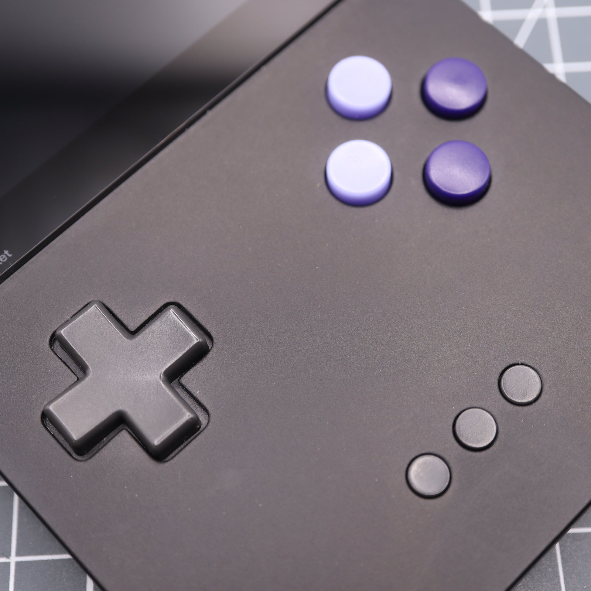 A close-up of the Analogue Pocket - Custom Button - Famicon Style handheld gaming console highlights its custom-cast buttons: a black D-pad, purple A/B buttons, and three small black menu buttons on a matte black surface with part of the screen visible.