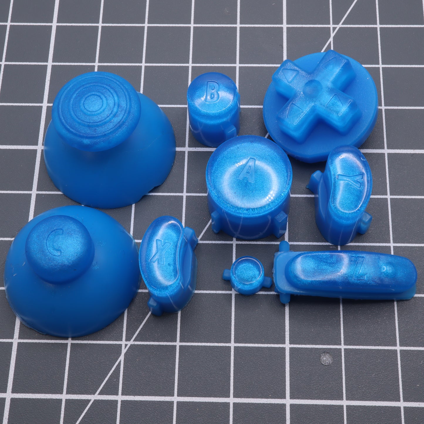 Assorted custom-cast GameCube - Custom Button - Blueberry Candy buttons with embossed letters and symbols on a gray grid background.