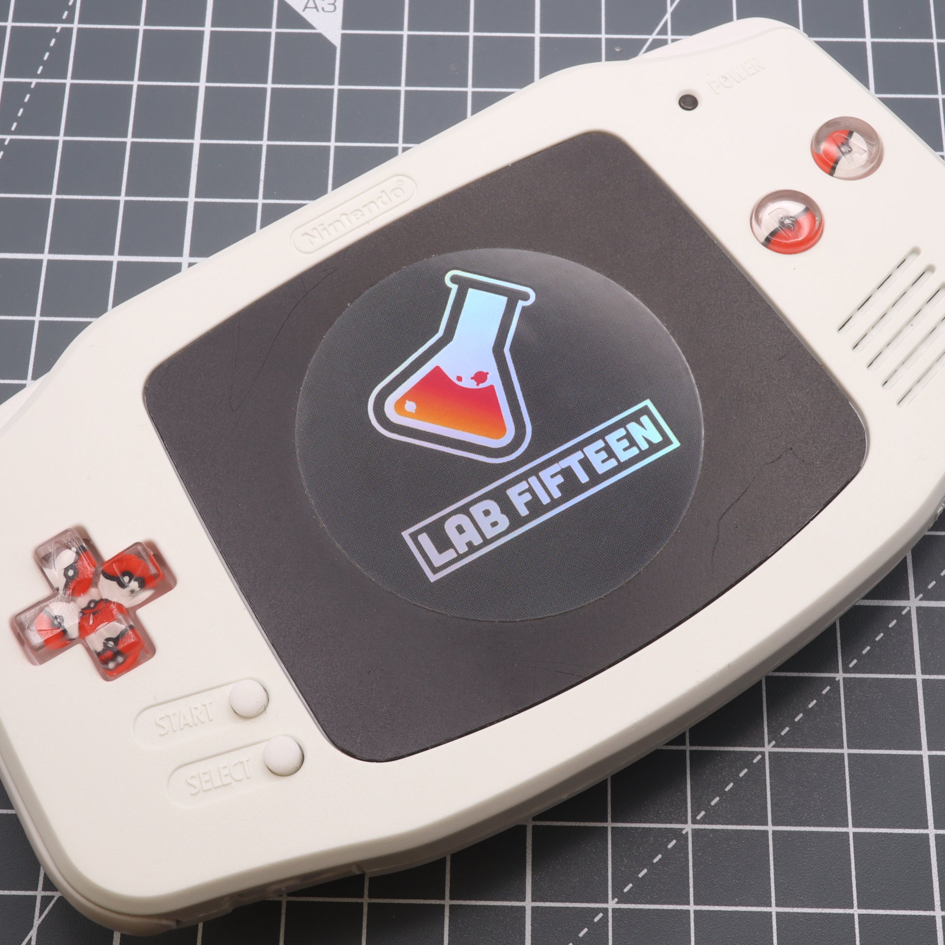 A white and black Game Boy Advance with labfifteen custom-cast Pokeball buttons.