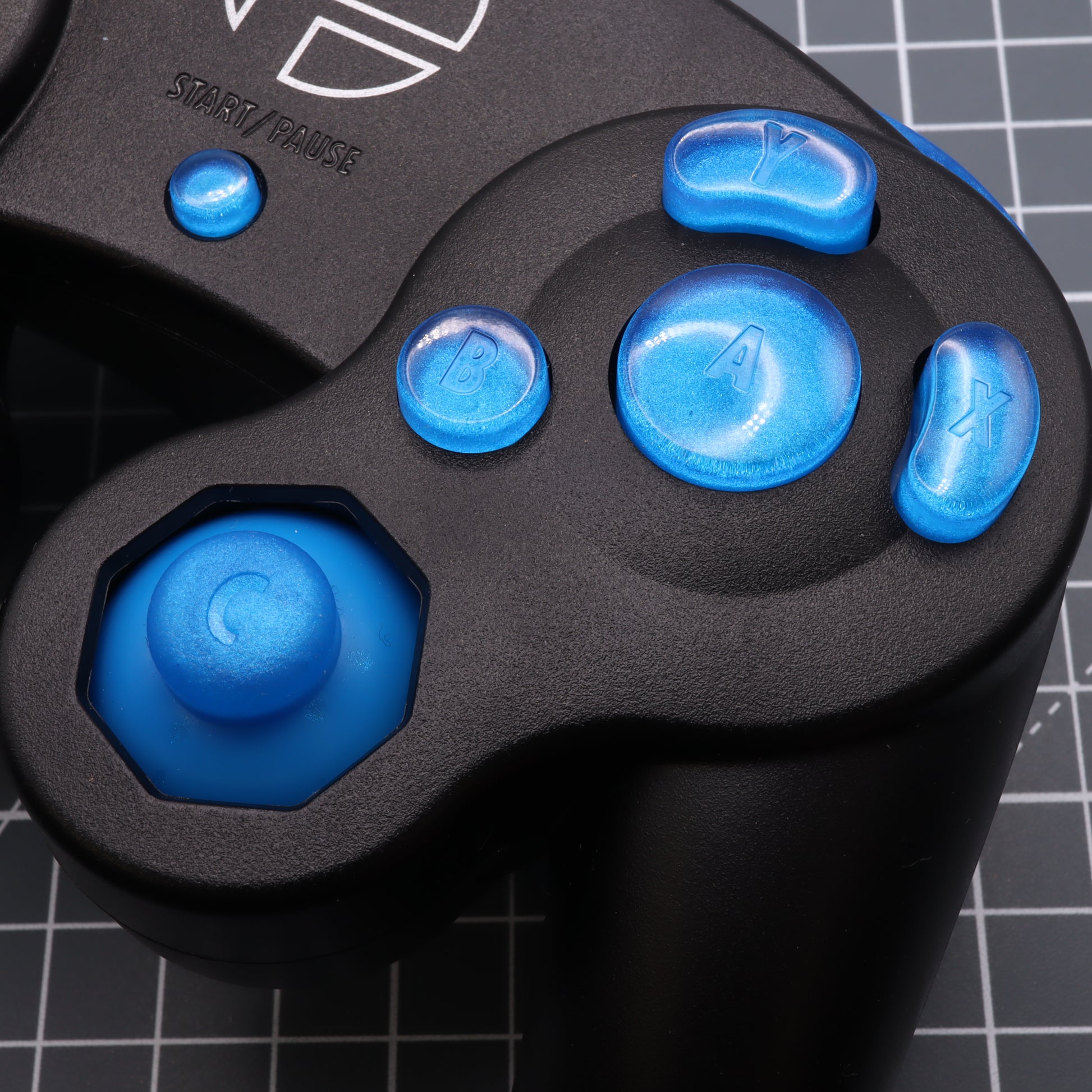 Close-up of a black GameCube controller with custom-cast blue buttons on a patterned surface.