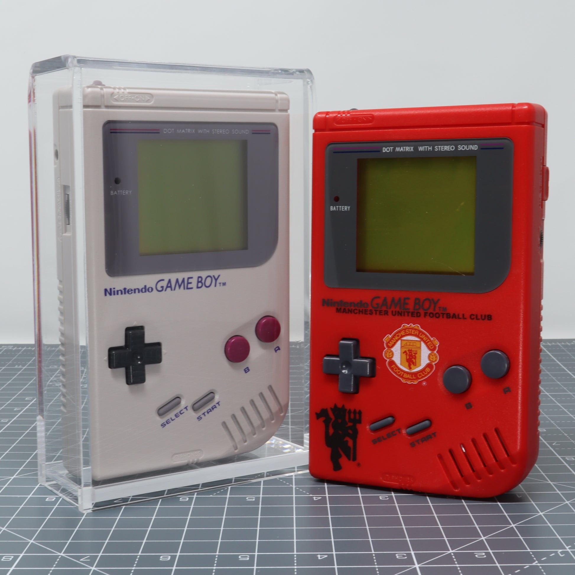 Two Game Boy DMG handheld consoles, one transparent and the other in Manchester United football club colors, are displayed side by side in acrylic display capsules.